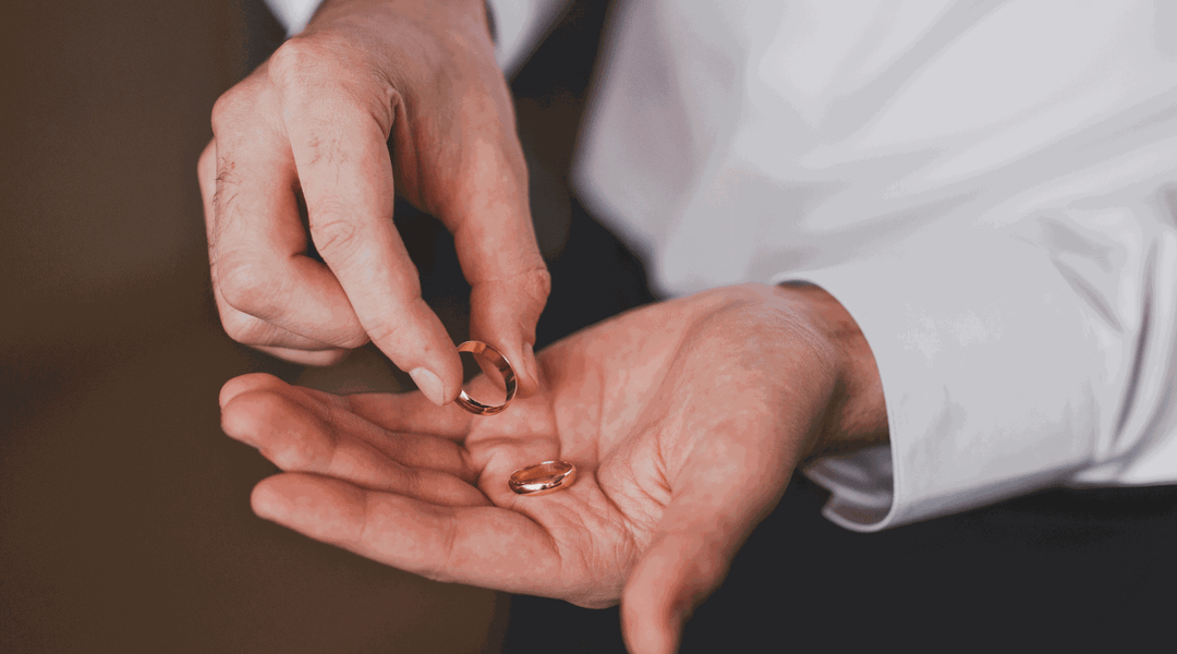 A man in a white shirt drops one gold wedding band into his open palm, which already holds a second matching band