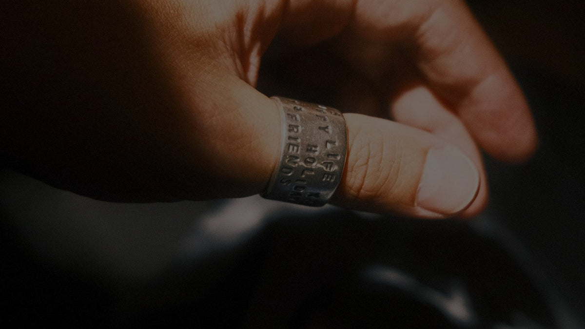 Close-up of a hand with a wide letter-stamped metal band on the ring finger, lit low against a dark background