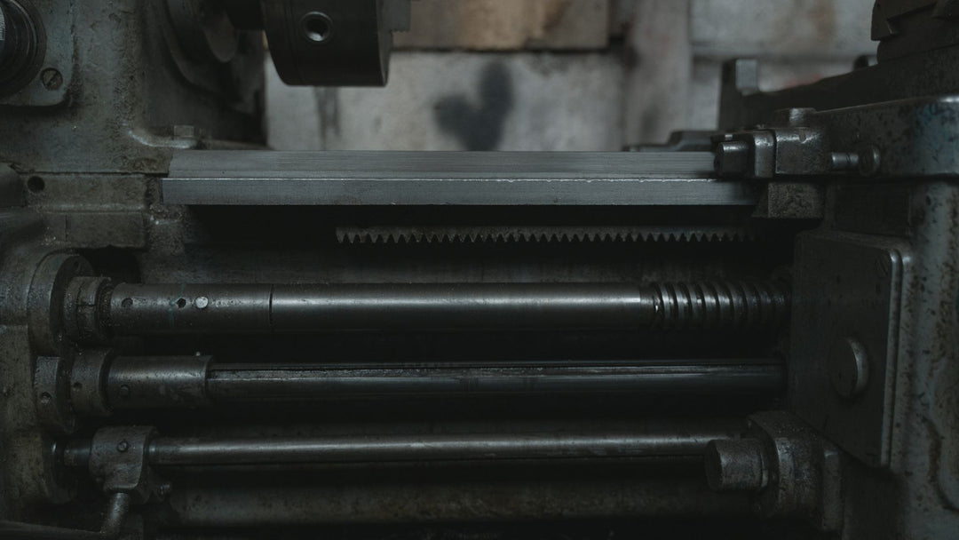 Close macro of a dark industrial lathe's gear rack, leadscrew, and tailstock mechanism, worn gunmetal-gray metal with visible tool marks, no people in frame.