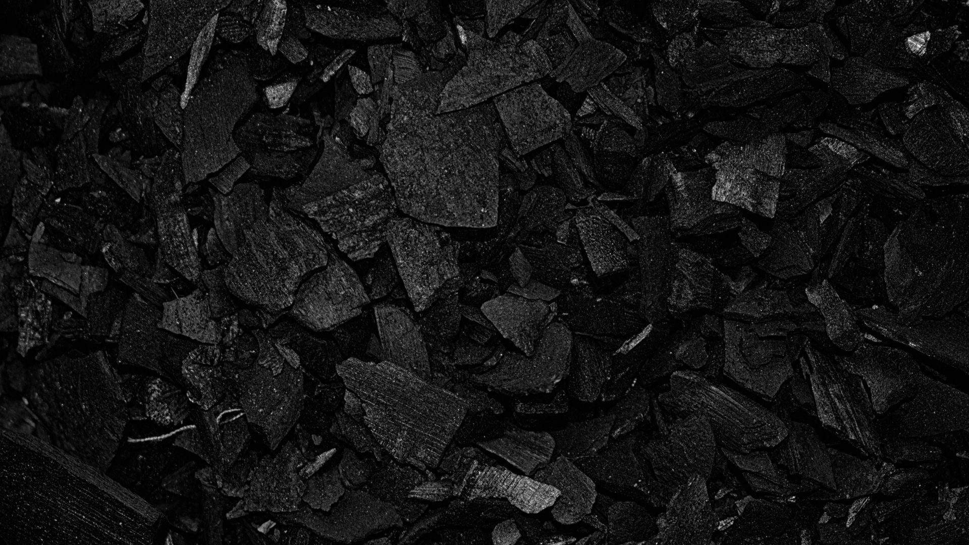 An overhead field of broken black charcoal chunks filling the frame, matte faces with fine pale grain catching a soft top light