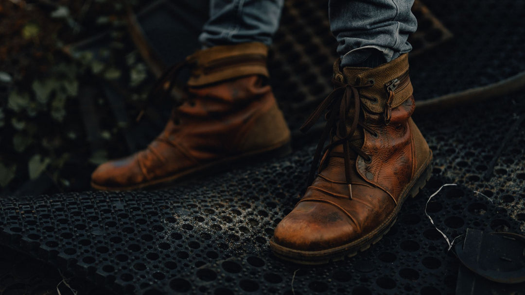 Close-up of a man's feet in worn brown leather lace-up boots standing on a dark metal grate, jeans visible above the boots