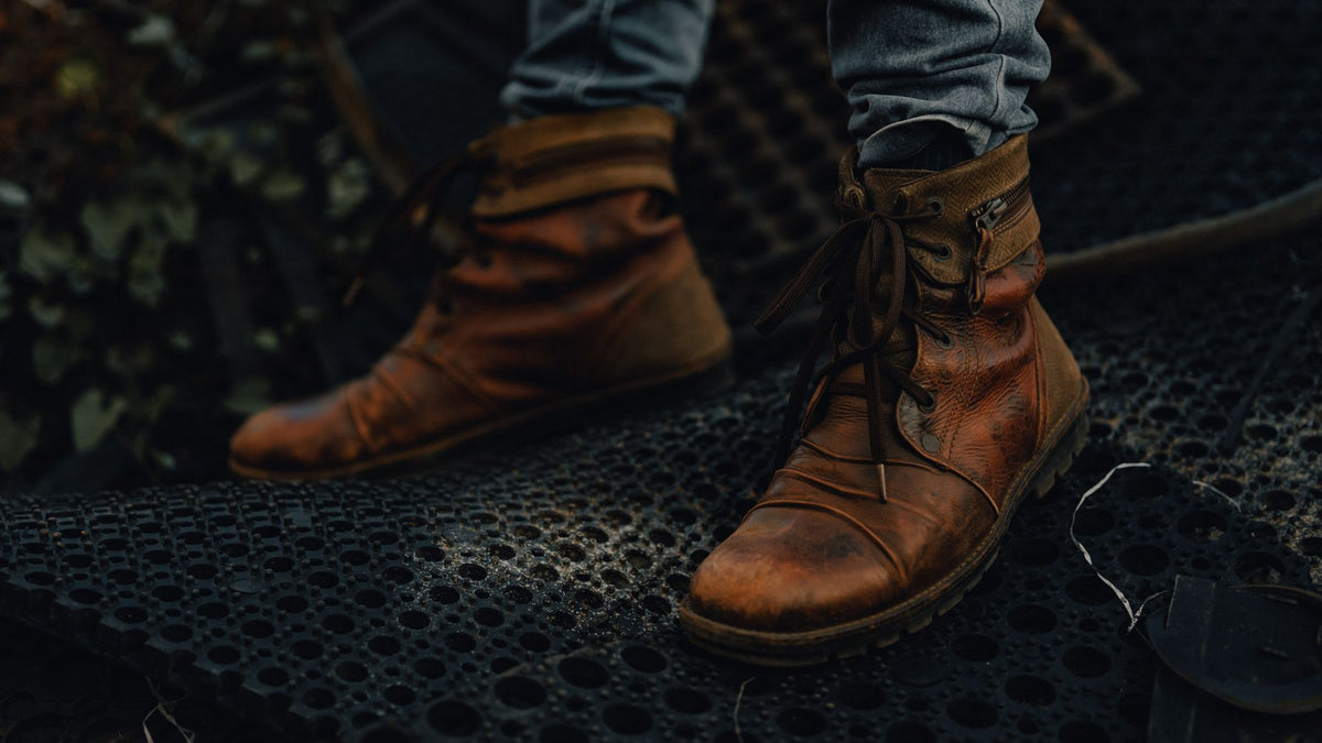 Close-up of a man's feet in worn brown leather lace-up boots standing on a dark metal grate, jeans visible above the boots