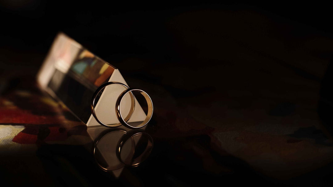 Two plain gold wedding bands leaning against a triangular glass prism on a dark reflective surface