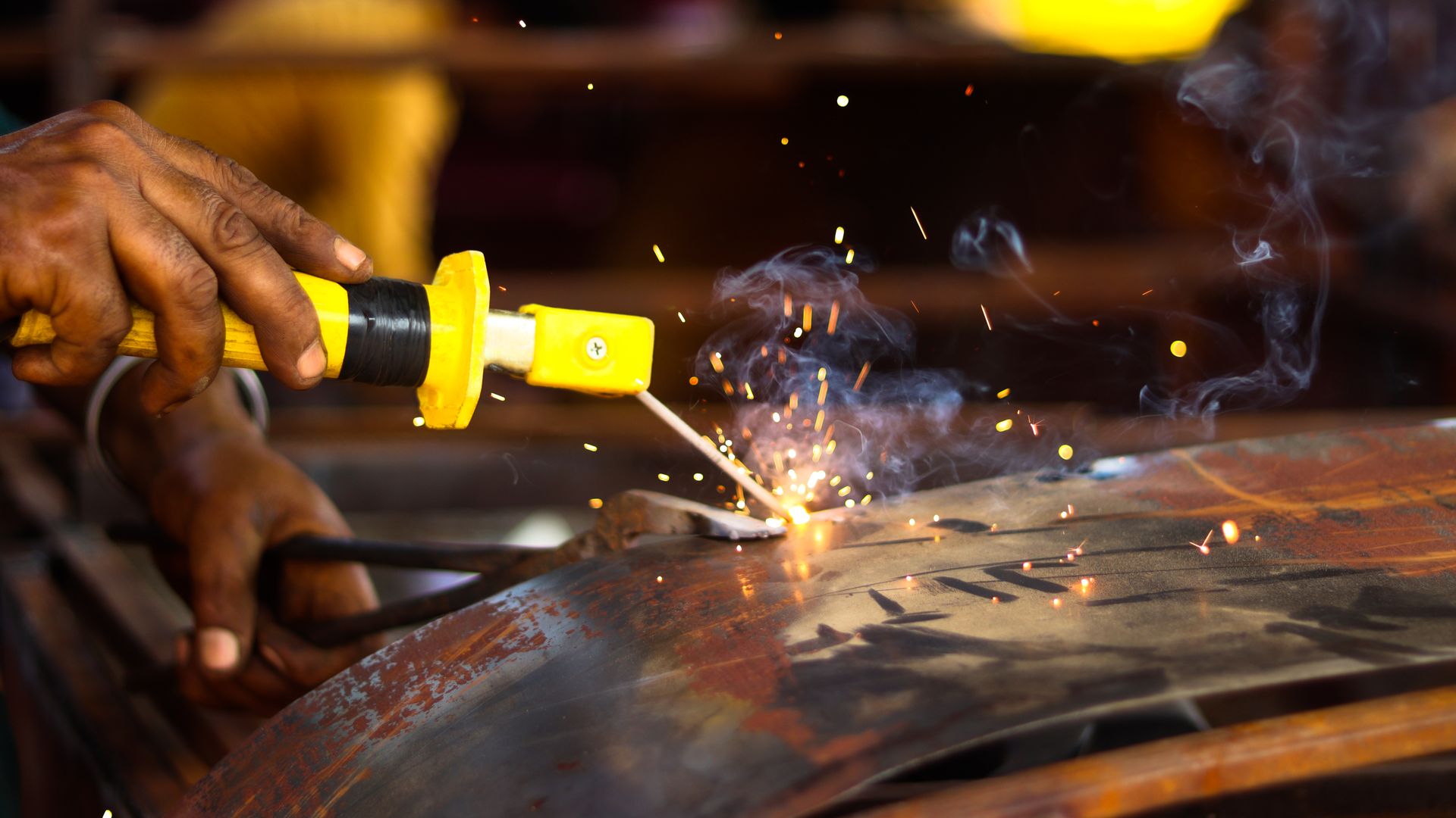 A hand running a yellow-handled cutting torch along rusted curved sheet steel, orange sparks and pale smoke rising in a dark workshop