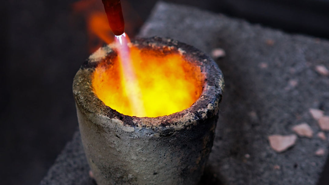 A jeweler's torch flame playing over molten metal glowing yellow inside a small ceramic crucible on a dark stone slab