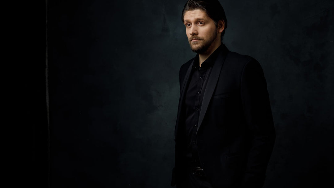 A bearded man in a black suit and black shirt standing against a dark studio background