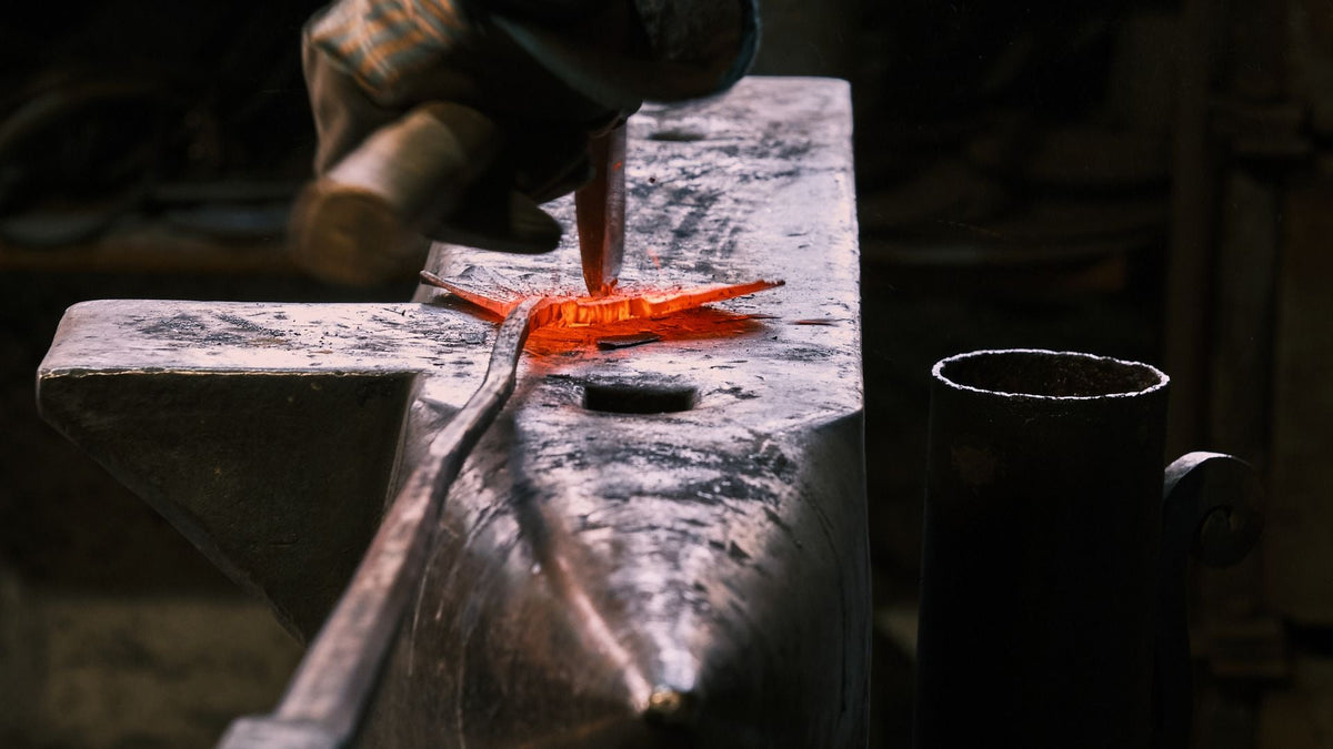 A glowing red-hot steel bar being cut with a chisel on a scarred anvil in a dark workshop