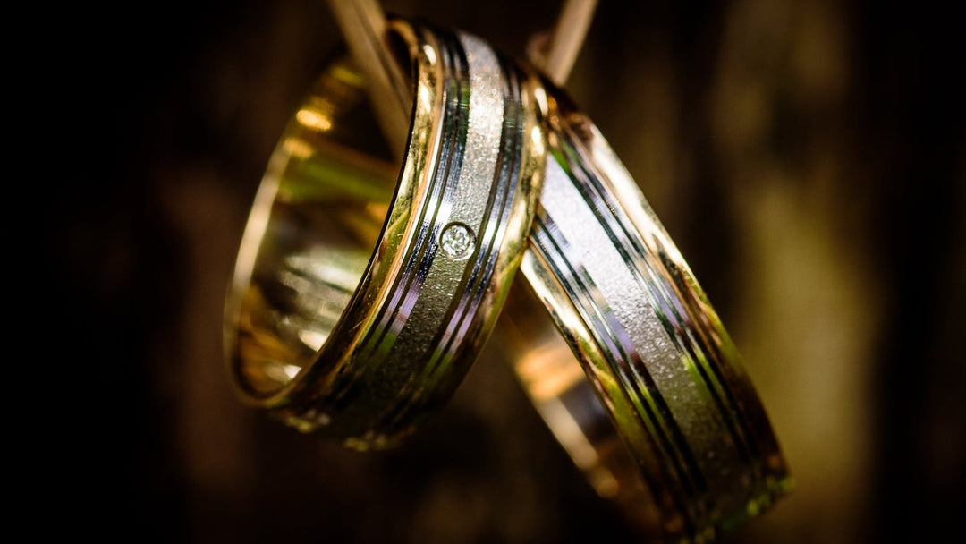engagement ring vs wedding ring for men — two two-tone wedding bands hanging on dark string, moody close-up — FoundryCut