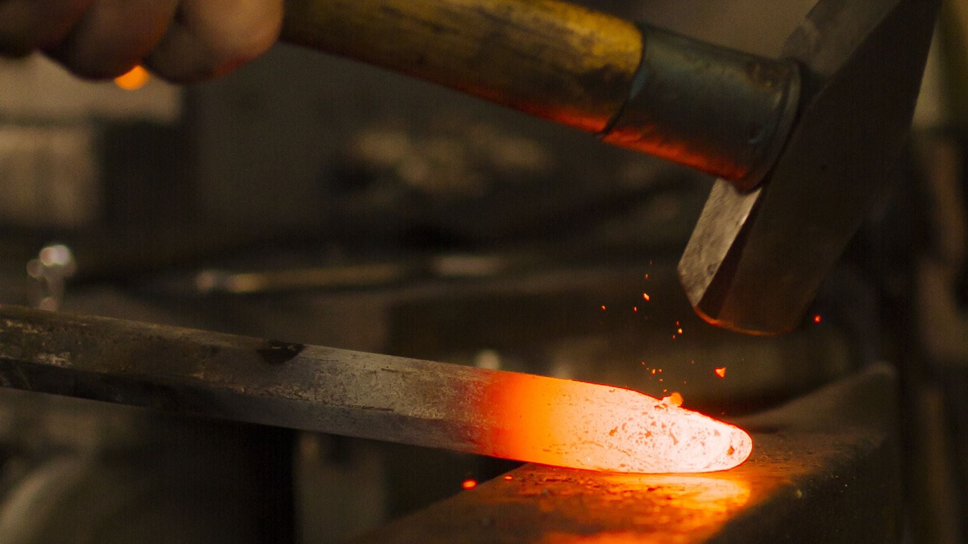 A hammer head striking a bar of steel glowing white-orange at the contact point on a dark anvil, with sparks lifting off