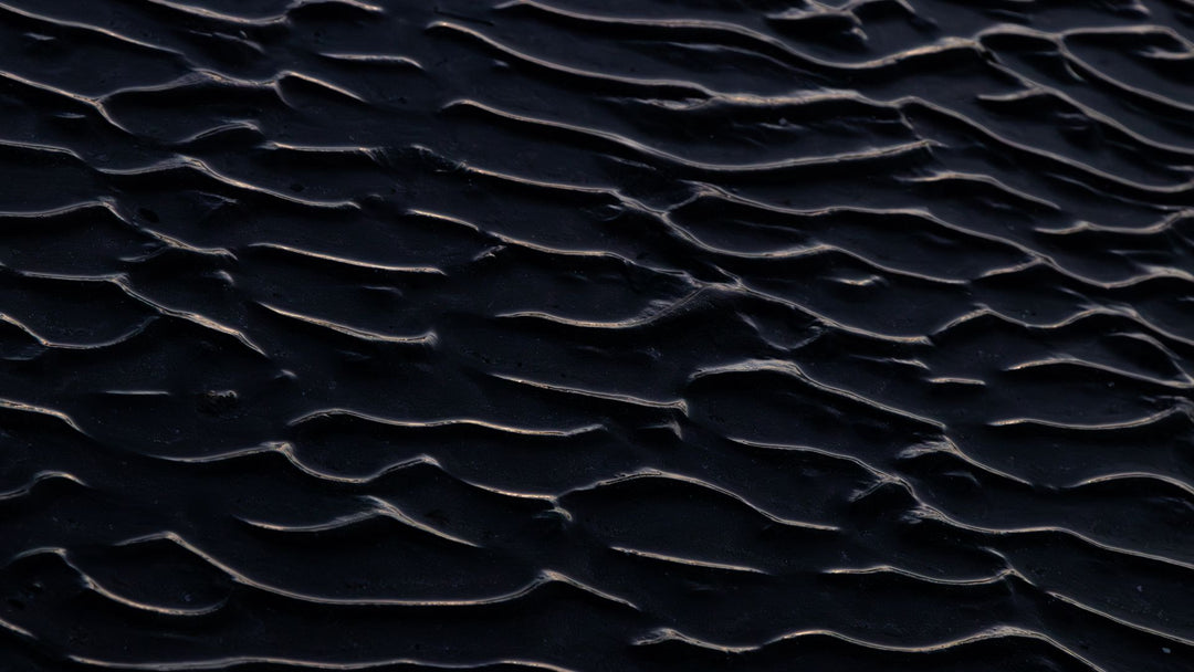 Overhead view of dark wet sand ripples, each crest catching a thin pale highlight against near-black troughs