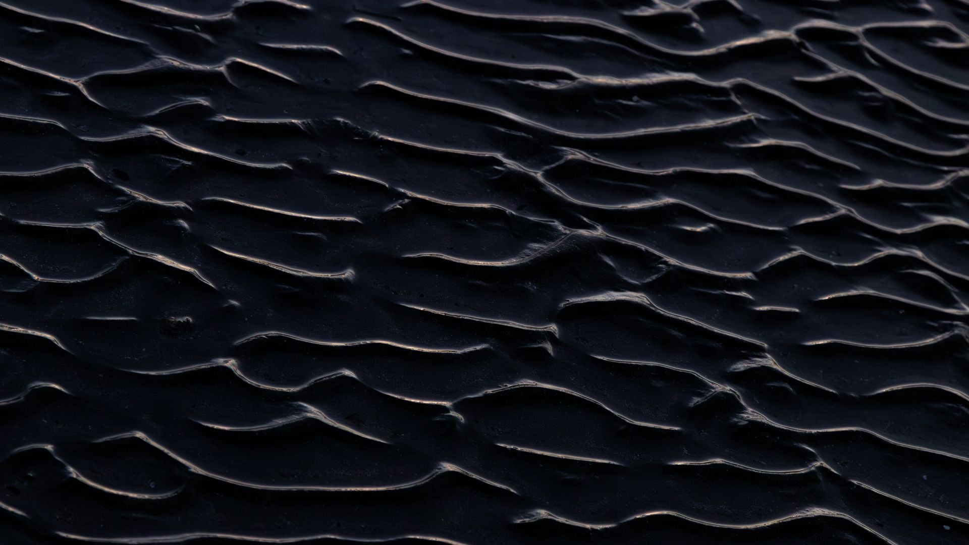 Overhead view of dark wet sand ripples, each crest catching a thin pale highlight against near-black troughs