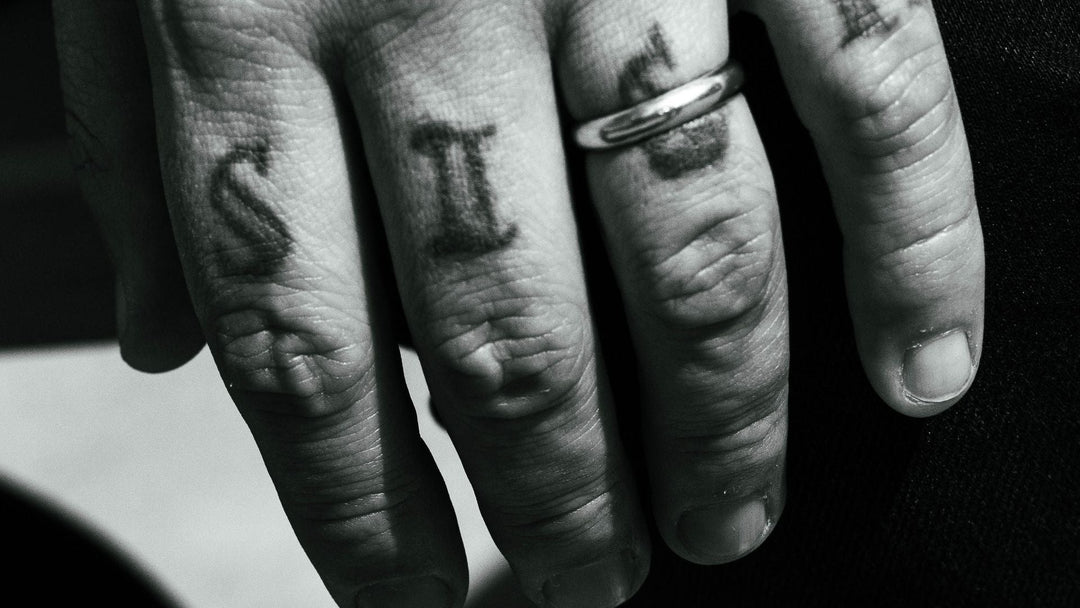 black and white close-up of a man's hand with knuckle letter tattoos and a plain metal band on the ring finger, FoundryCut