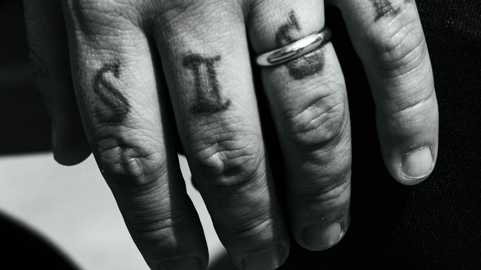 black and white close-up of a man's hand with knuckle letter tattoos and a plain metal band on the ring finger, FoundryCut