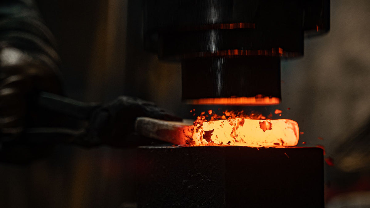 A blacksmith's gloved hands holding glowing orange metal under a power hammer as sparks scatter in a dark forge
