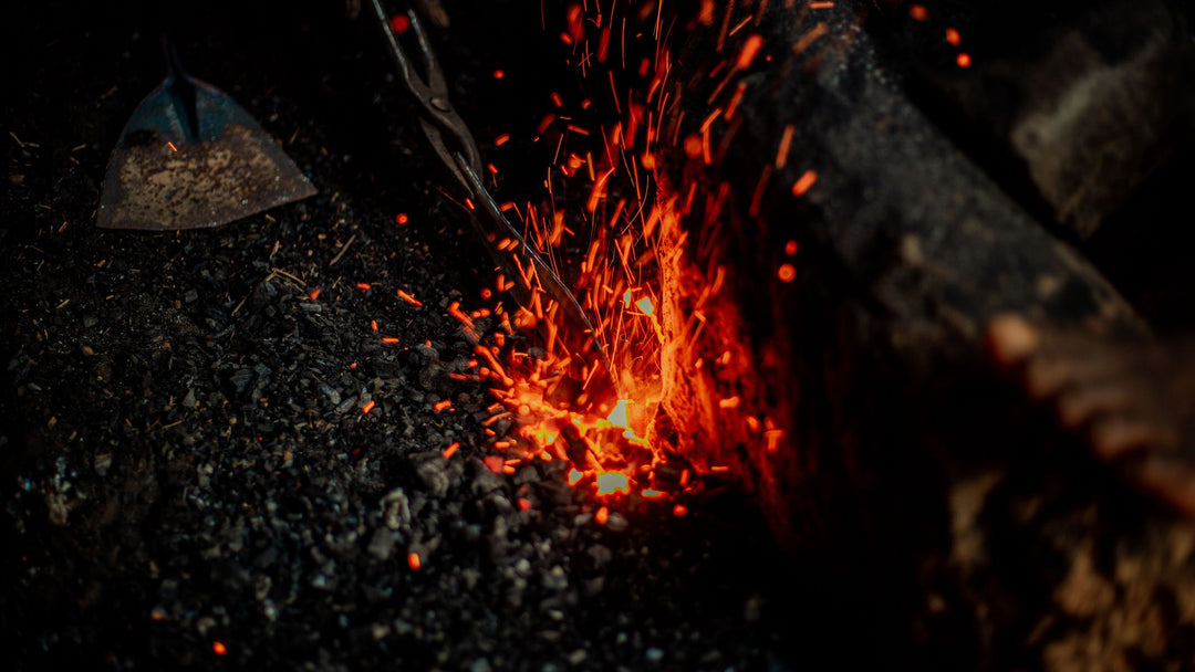 Glowing orange embers and sparks rising from a bed of black forge coal, with metal tongs entering the frame from above