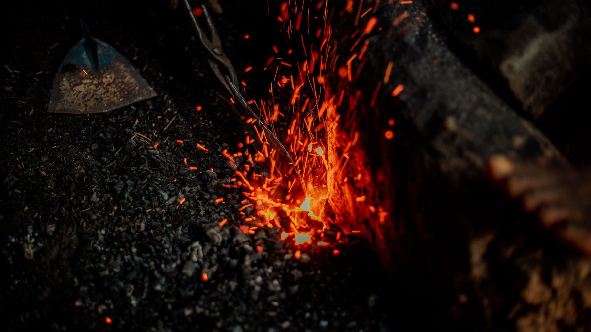 Glowing orange embers and sparks rising from a bed of black forge coal, with metal tongs entering the frame from above