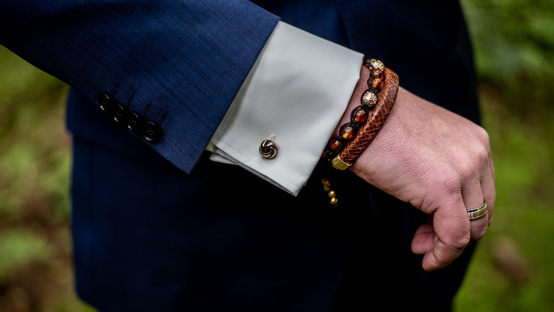 A man's hand and wrist in a navy suit sleeve, wearing a thin wedding band, a knot cufflink, and two beaded bracelets, with greenery blurred behind him