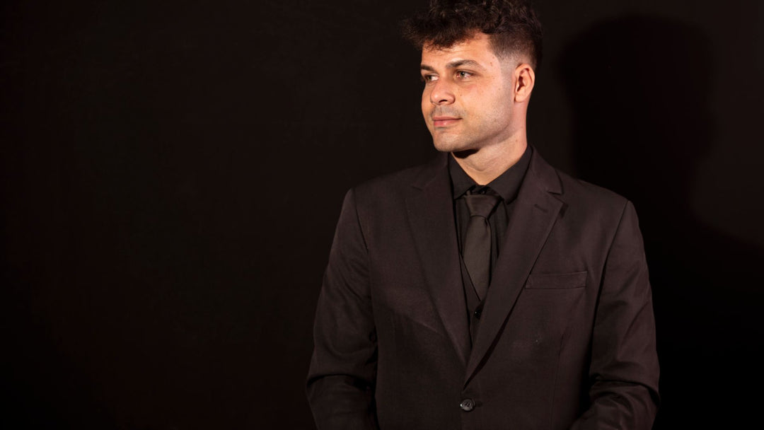 A man in a black suit, black shirt and black tie standing alone against a dark studio wall