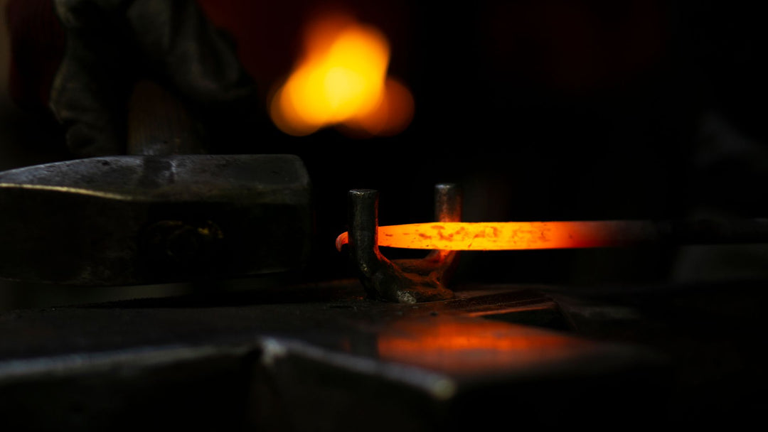 A bar of steel glowing orange held in a forging jig on a dark anvil, a blacksmith's hammer resting at the left and a soft forge flame out of focus behind