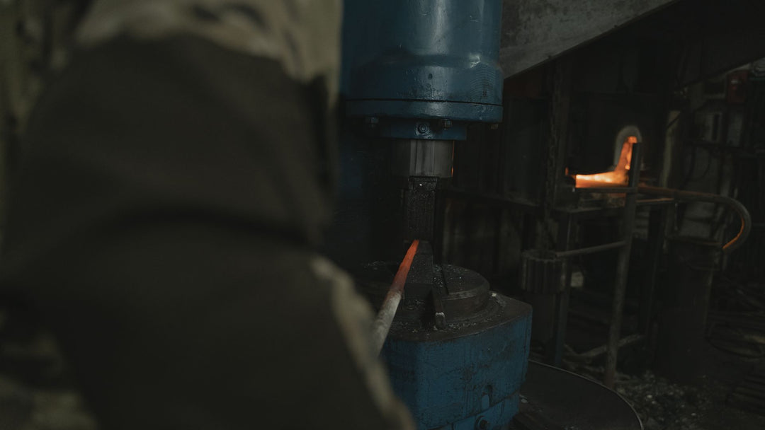 Blue industrial power hammer pressing a glowing orange steel bar against its die inside a dark forge, with a second heated piece resting in a rack to the right