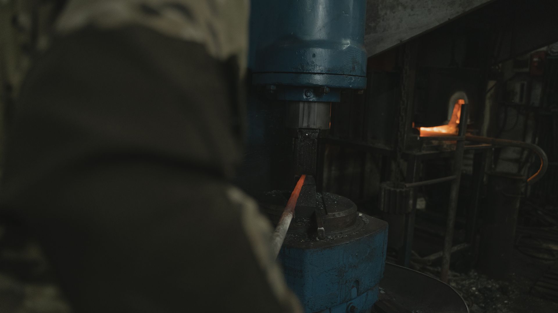 Blue industrial power hammer pressing a glowing orange steel bar against its die inside a dark forge, with a second heated piece resting in a rack to the right
