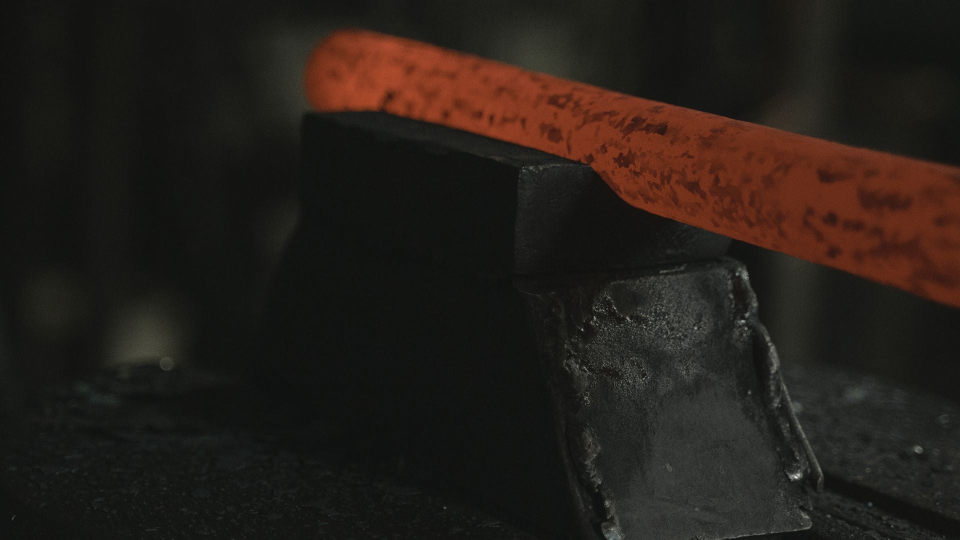 A flat bar of steel glowing red-orange laid diagonally across a black anvil block in a dark forge workshop