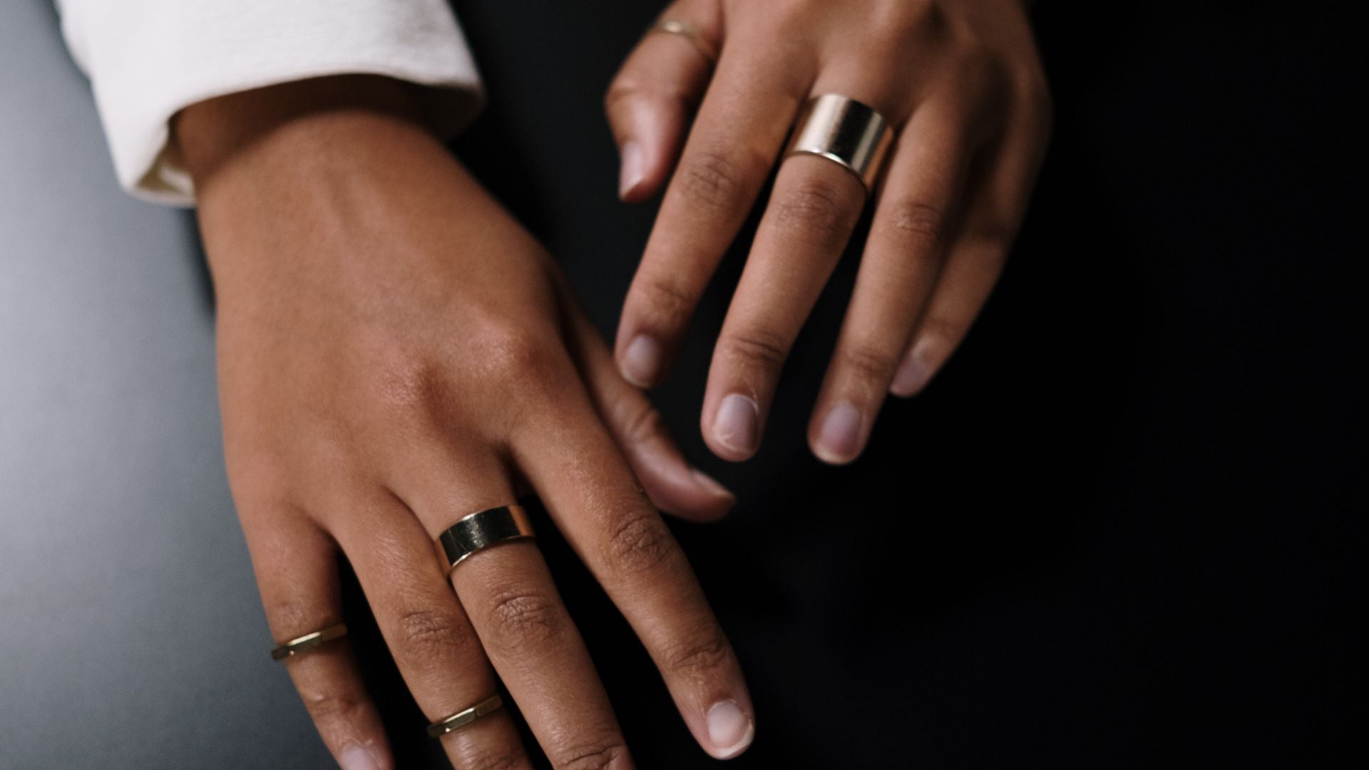 Two hands resting on a dark surface wearing five plain metal rings across different fingers