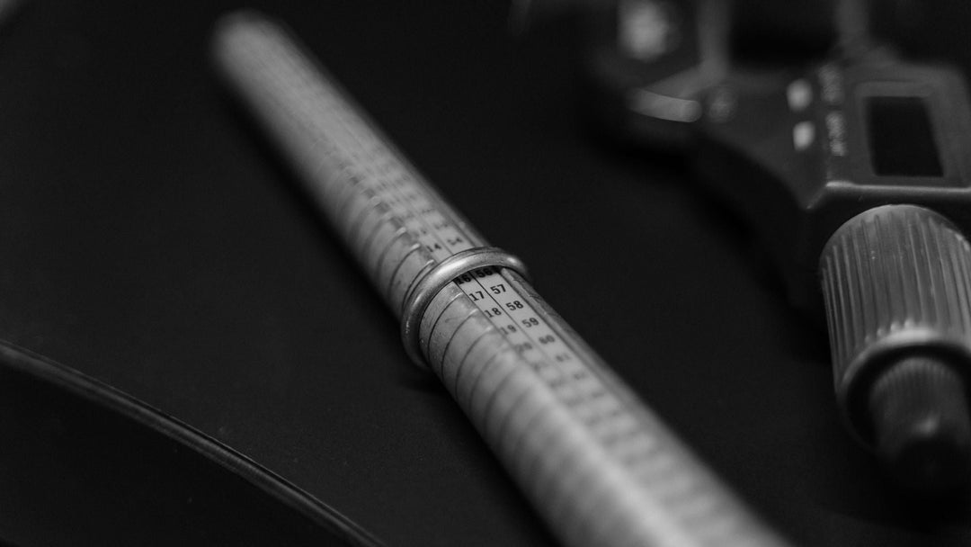 A wedding band resting on a numbered ring-sizing gauge beside a caliper, shot in near-black and white
