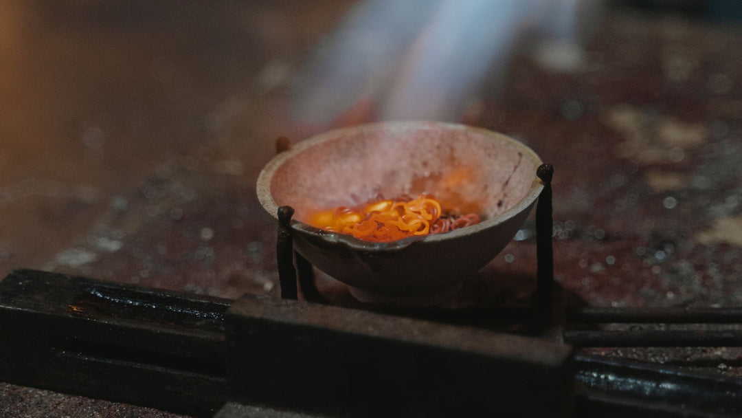 A jeweler's torch flame playing over molten metal glowing yellow inside a small ceramic crucible on a dark stone slab