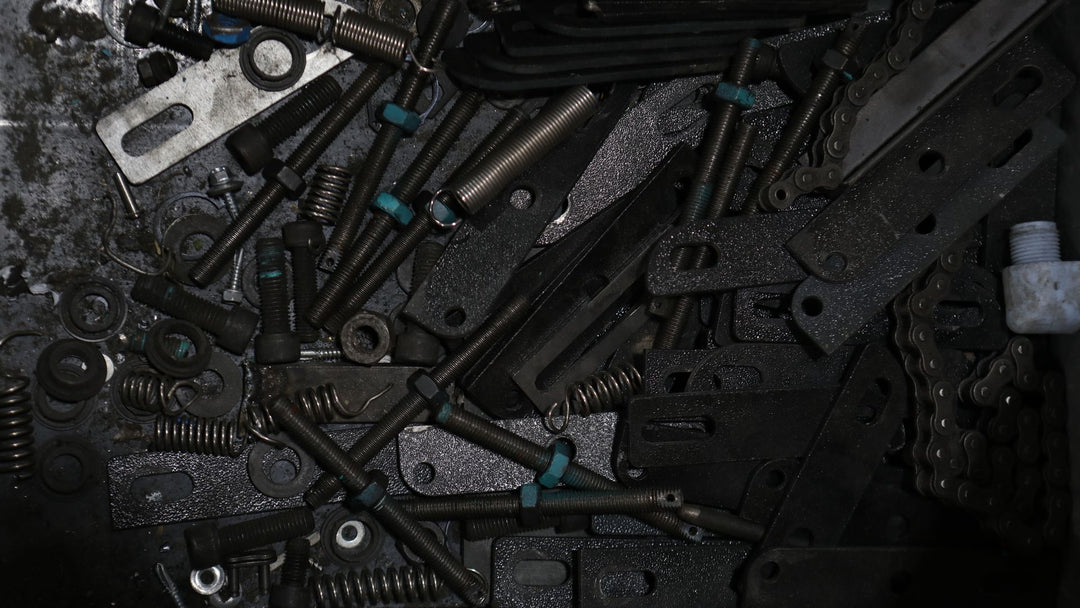 An overhead spread of loose workshop hardware on a dark oiled surface: threaded bolts, coil springs, steel brackets and a length of roller chain