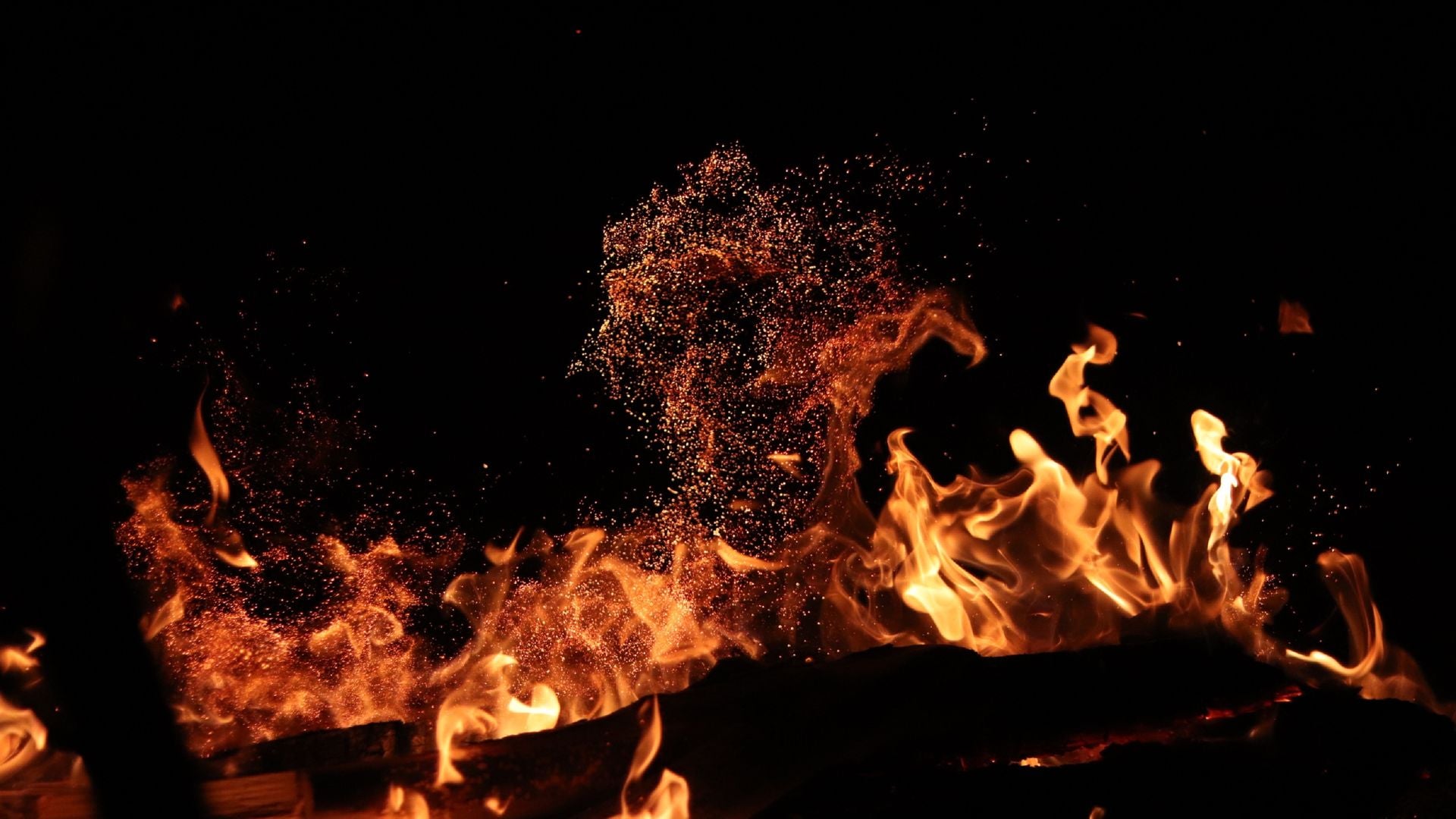 Orange flame tongues and a rising column of embers against a black night field, with a dark log silhouetted along the bottom edge