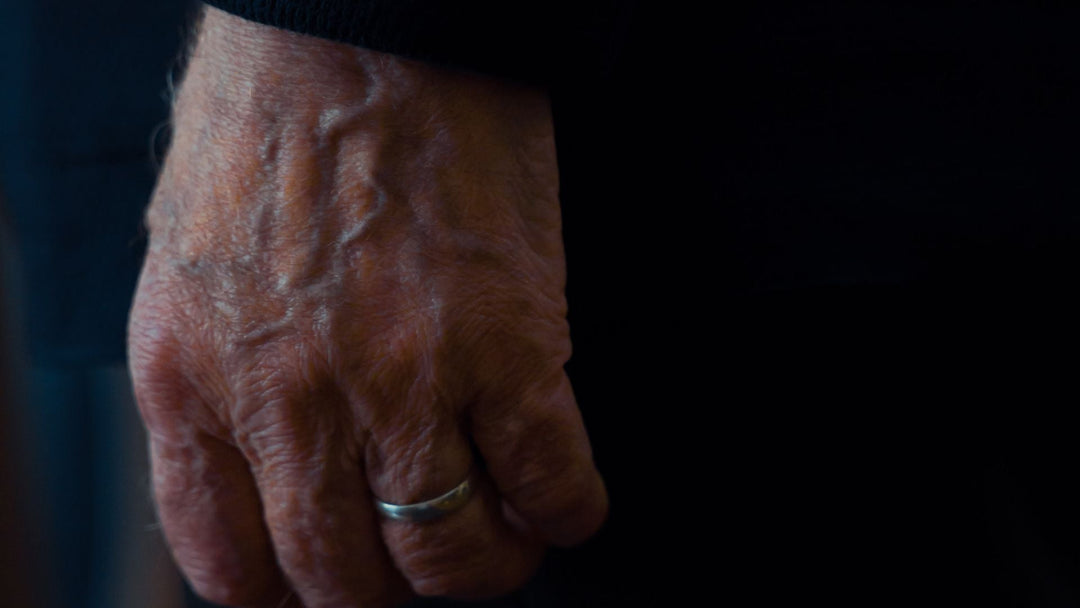 A weathered older man's hand wearing a plain worn wedding band on the ring finger against a near-black background