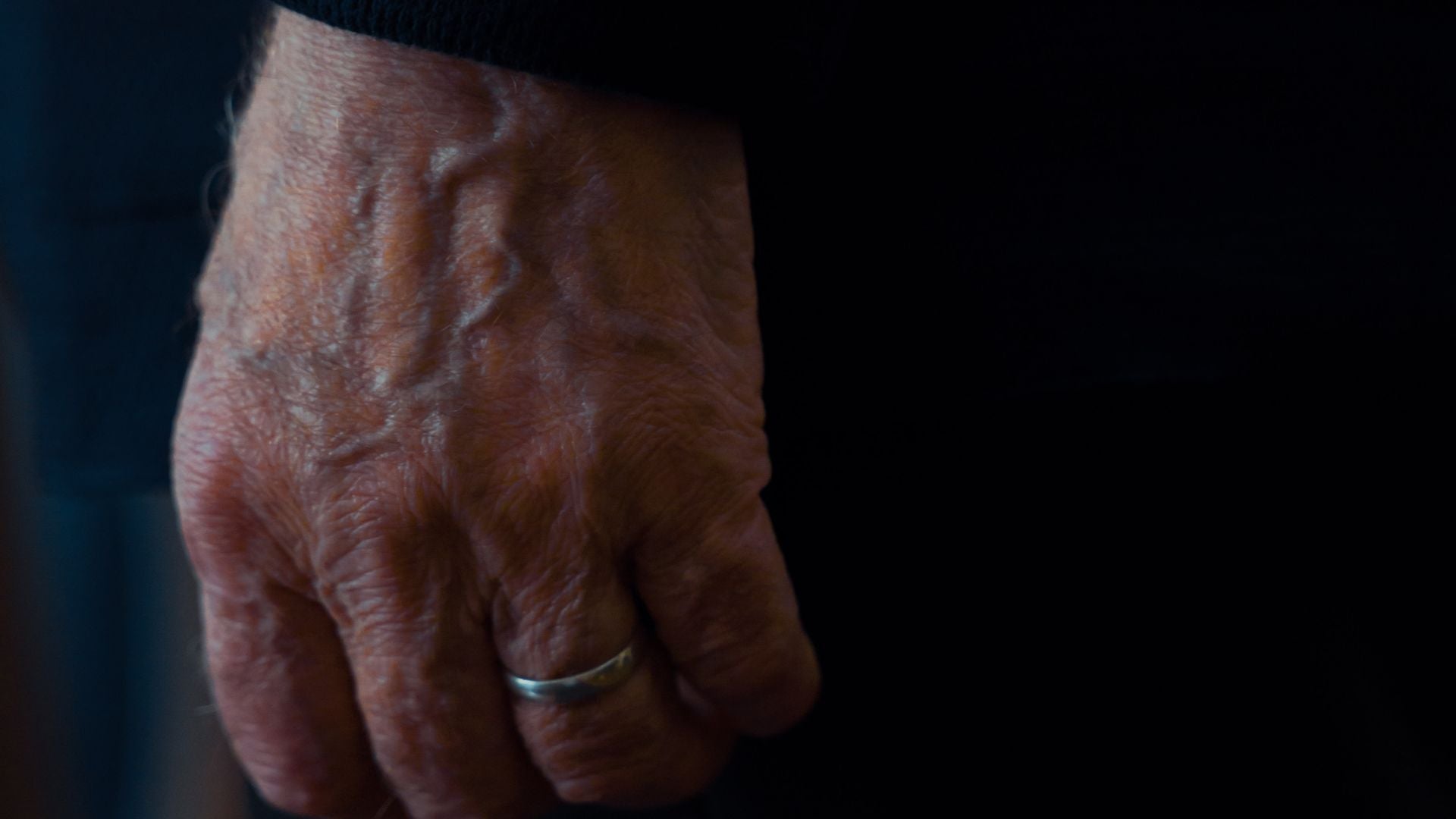 A weathered older man's hand wearing a plain worn wedding band on the ring finger against a near-black background