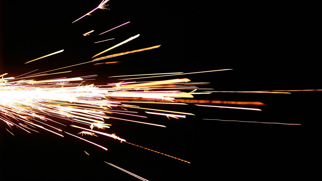 A long-exposure burst of white-gold grinder sparks streaking across a black field, the dense ignition point at the left edge