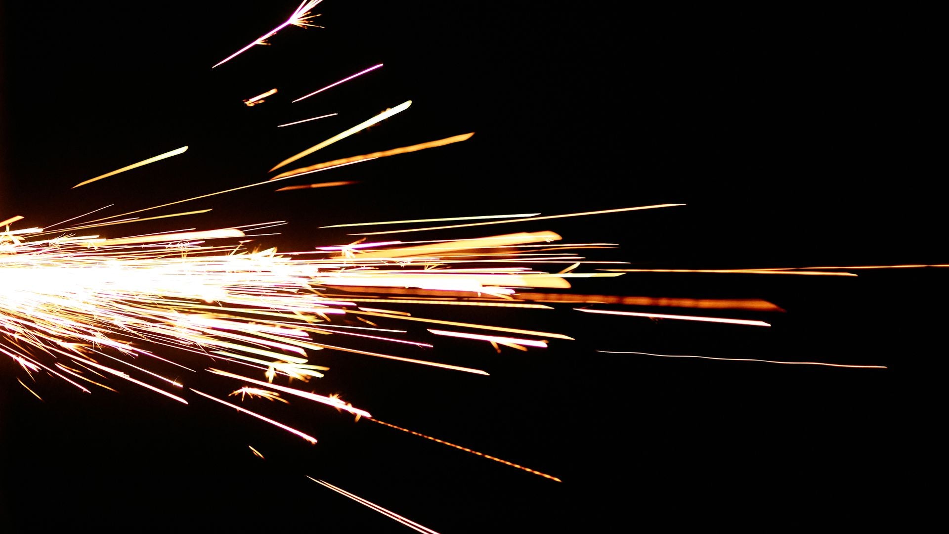 A long-exposure burst of white-gold grinder sparks streaking across a black field, the dense ignition point at the left edge