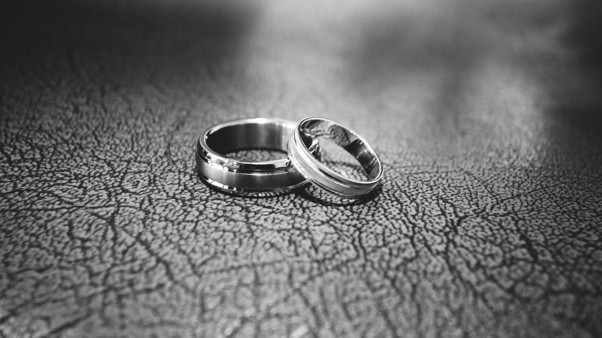 Black-and-white photo of two metal wedding bands leaning together on a cracked leather surface
