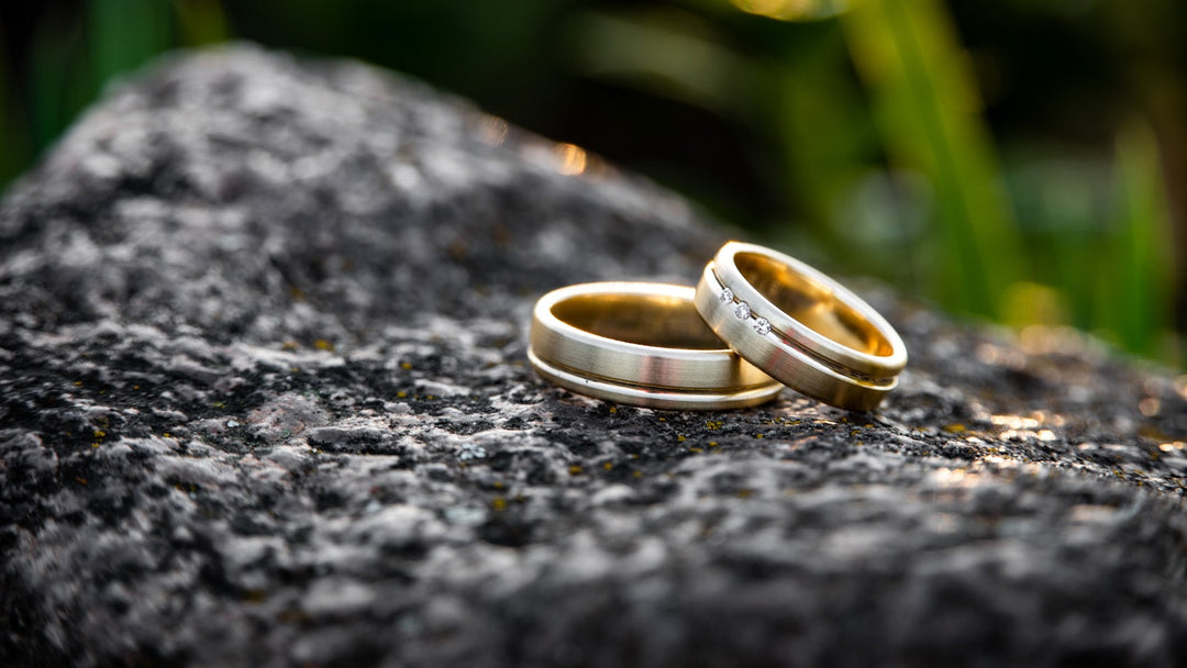 Two brushed gold wedding bands, one set with three small diamonds, resting together on dark speckled granite with greenery behind