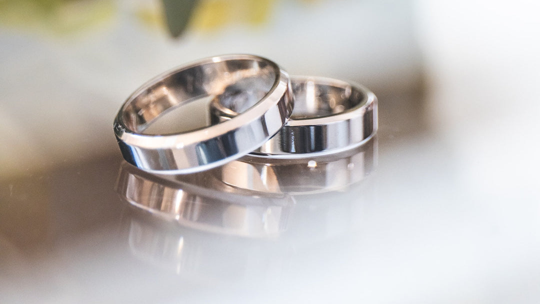 Close-up of two polished silver-tone wedding bands with stepped beveled edges resting on a reflective surface
