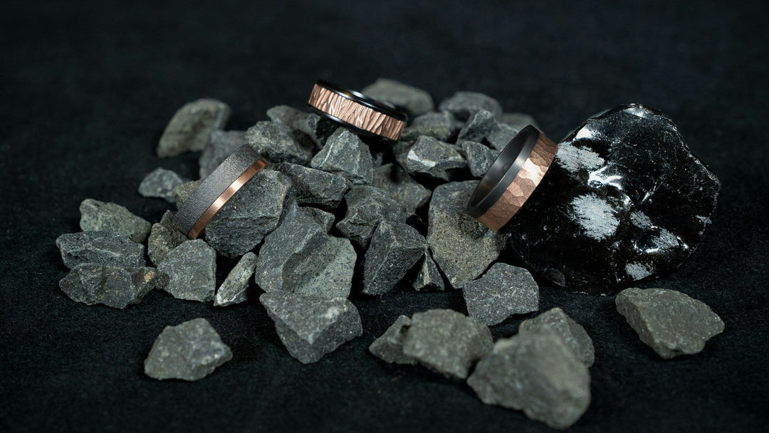 Three men's two-tone tungsten wedding bands arranged on dark crushed gravel beside a piece of obsidian